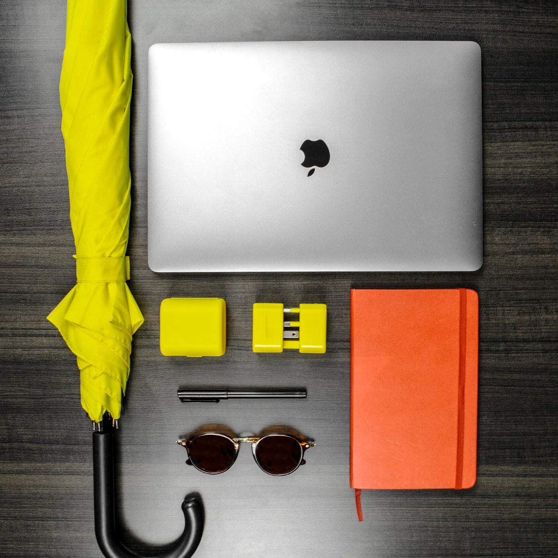 The universal travel adapter along with an umbrella, a pen, sunglasses, notebook and MacBook Pro laptop. The travel adapter is the World Traveler Jetsetter universal travel adapter