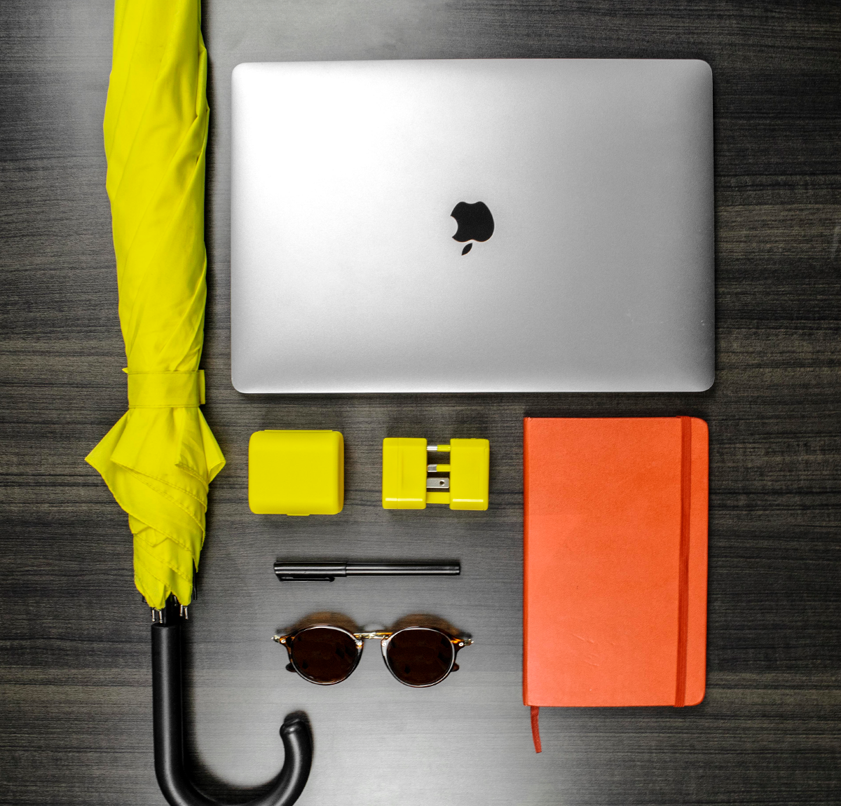 The universal travel adapter along with an umbrella, a pen, sunglasses, notebook and MacBook Pro laptop. The travel adapter is the World Traveler Jetsetter universal travel adapter